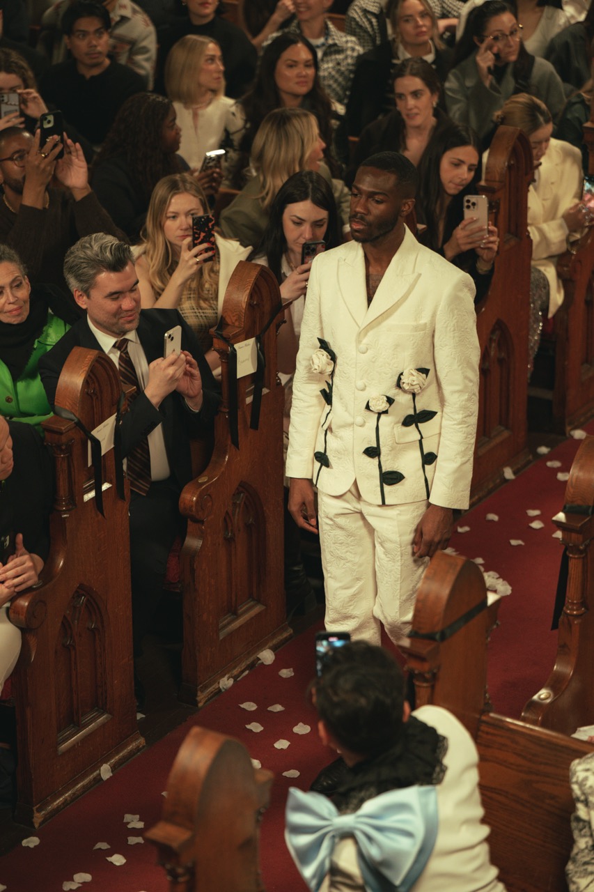 Model walks down the aisle in a white floral-embellished suit during Tanner Fletcher's immersive bridal fashion show at New York Bridal Fashion Week, as guests watch from the church pews and photograph the runway.